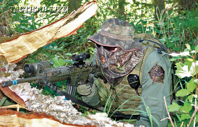 An AST SERT countersniper decked in camouflage holds a perimeter overwatch position from concealment during a day-long hostage-rescue training operation.