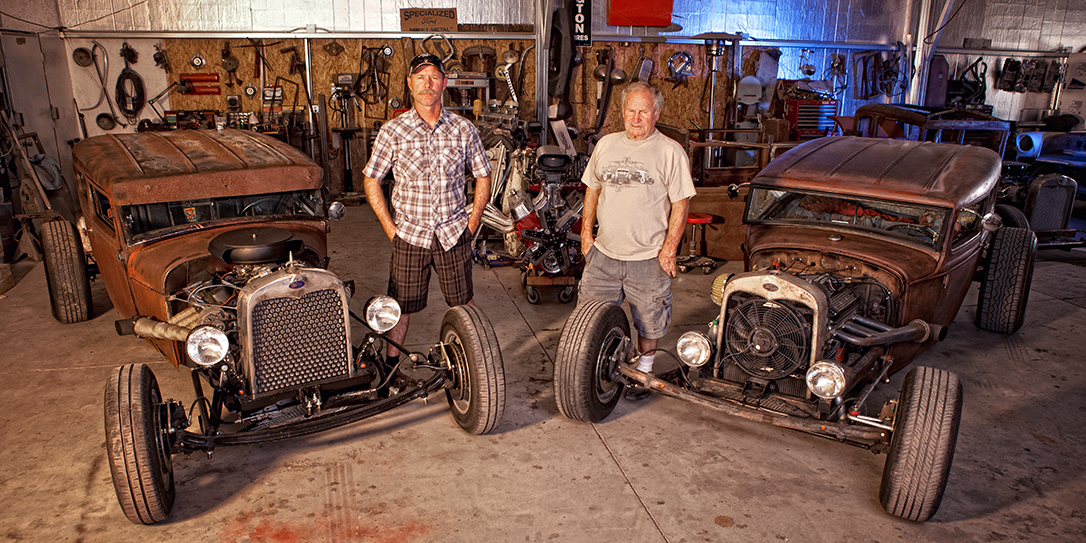 Two proud men standing with their rat rod creations in there backyard garage.